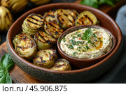 An appetizing dish presented in a ceramic bowl. In the center of the plate is a creamy sauce, garnished with fresh herbs (such as basil) and olive oil, and lightly sprinkled with spices (paprika or pepper). Around the sauce are pieces of baked artichokes with appetizing grill stripes. The dish looks fresh and flavorful, which emphasizes its ideal for a light snack or appetizer. Green leaves are visible in the background, adding naturalness and aesthetics to the composition. Стоковое AI-изображение, AI-художник Анна Кривицкая / Фотобанк Лори