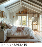 A cozy bedroom in the Provence style, located on the attic floor. The interior is made in light pastel colors, which creates a feeling of warmth and comfort. The main attention is drawn to a large bed with a high carved headboard, covered with a white down blanket with a delicate floral pattern. Soft pillows and a blanket are neatly laid out on the bed.The room has a lot of natural light, which penetrates through large windows with transparent curtains. The floor is made of natural wood with warm shades, which emphasizes the atmosphere of a country house. On the bedside table there is a vase of flowers, adding freshness. Above the bed on the wall hang paintings with landscapes that are in harmony with the overall style. On the shelves and chest of drawers there are wicker baskets and decorative elements, giving the room completeness and comfort. Стоковое AI-изображение, AI-художник Анна Кривицкая / Фотобанк Лори
