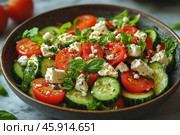 A close-up of a delicious Greek salad served in a deep ceramic bowl. The salad contains fresh vegetables: sliced ​​cucumbers, halves of juicy cherry tomatoes and basil leaves. The central place is occupied by pieces of soft white feta cheese, which are slightly crumbled, creating a textural contrast. The salad is dressed with olive oil, and freshly ground black pepper is added to add a piquant aroma. The light marble surface of the table in the background emphasizes the richness of the colors of the dish, and nearby you can see the blurred silhouettes of fresh tomatoes and greens. Стоковое AI-изображение, AI-художник Анна Кривицкая / Фотобанк Лори