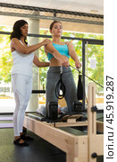 Focused Latin female instructor helping girl practicing pilates stretching exercises on reformer at gym. Individual coach. Стоковое фото, фотограф Яков Филимонов / Фотобанк Лори