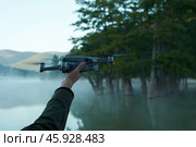 Outstretched arm with quadrocopter on the background of a picturesque morning lake in the mountains with a grove of cypresses in a misty haze. Copy space. Стоковое фото, фотограф Роман Дзюбало / Фотобанк Лори