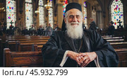 Smiling old christian priest sitting inside church. Стоковое AI-видео, аниматор Александр Довянский / Фотобанк Лори