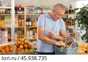 Elderly man with paper list in hands, walks around minimarket, puts groceries in market basket. Стоковое фото, фотограф Яков Филимонов / Фотобанк Лори