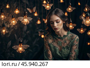 Portrait of a beautiful girl in a green dress among many suspended light bulbs and butterflies fluttering around her. Daydreaming. A magical atmosphere. Стоковое AI-изображение, AI-художник Photojuli / Фотобанк Лори