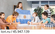 Group of curious preteen learners and young female teacher with flag of Argentina in classroom of academy. Стоковое фото, фотограф Яков Филимонов / Фотобанк Лори