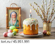 Orthodox Easter. Easter cake, painted eggs, icons, faces of saints, flowers, church candles and Easter decor on a wooden table in an old village house. Стоковое AI-изображение, AI-художник Photojuli / Фотобанк Лори