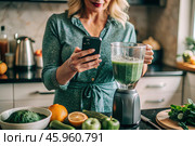 An elderly woman in the kitchen is looking at healthy food recipes on her phone and preparing a vegetable smoothie made from broccoli in a blender. Healthy detox diet drink. Стоковое AI-изображение, AI-художник Photojuli / Фотобанк Лори