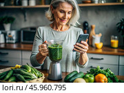 An elderly woman in the kitchen is looking at healthy food recipes on her phone and preparing a vegetable smoothie made from broccoli in a blender. Healthy detox diet drink. Стоковое AI-изображение, AI-художник Photojuli / Фотобанк Лори