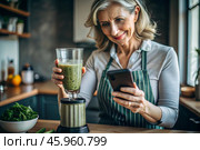 An elderly woman in the kitchen is looking at healthy food recipes on her phone and preparing a vegetable smoothie made from broccoli in a blender. Healthy detox diet drink. Стоковое AI-изображение, AI-художник Photojuli / Фотобанк Лори