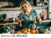 An elderly woman in the kitchen is looking at healthy food recipes on her phone and preparing a vegetable smoothie made from broccoli in a blender. Healthy detox diet drink. Стоковое AI-изображение, AI-художник Photojuli / Фотобанк Лори