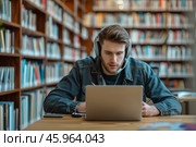 Handsome student focused on writing an essay with laptop in a quiet library during afternoon study hours. Стоковое AI-изображение, AI-художник Sergey Grishin / Фотобанк Лори
