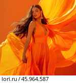 A young woman with long dark hair flowing freely in the wind. She is wearing a bright orange dress with a low neckline that also flutters, creating a sense of movement and drama. The dress is lightweight, made of flowing fabric reminiscent of silk or chiffon. The bright orange color of the dress harmonizes with the background, which is also in orange tones, enhancing the effect of sunlight and warmth. The woman wears large golden-orange earrings with decorative elements, emphasizing her elegance. Her facial expression is confident, and her pose is feminine and graceful. The overall atmosphere of the photo is dynamic, bright and stylish, reminiscent of a shoot for a fashion magazine or advertisement. Стоковое AI-изображение, AI-художник Анна Кривицкая / Фотобанк Лори