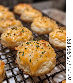 A close-up of a delicious baked good, cheese buns, placed on a metal rack. The buns have a golden crust with melted cheese on top. The cheese is topped with finely chopped herbs, likely parsley, which adds freshness and color contrast. The buns look soft and fluffy, with a delicate texture, and the cheese crust adds crunch. The background is slightly blurred, allowing the focus to be on the baked goods. The image creates a cozy home-cooking feel and perfectly conveys the aroma and texture of freshly baked goods. Стоковое AI-изображение, AI-художник Анна Кривицкая / Фотобанк Лори