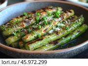 A dish of grilled asparagus served in a deep bowl. The asparagus has delicious grill marks that highlight its golden brown color and add texture. On top of the asparagus are fresh green peas, chopped herbs, and finely grated cheese that has melted slightly. A sprinkling of spices adds pops of color, such as red flakes (like chili peppers). The dish looks fresh and flavorful, thanks to the shiny butter that brings out the richness of the vegetables. The overall presentation is modern, and the background is blurred, which focuses the attention on the food. Стоковое AI-изображение, AI-художник Анна Кривицкая / Фотобанк Лори