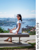 A 50-year-old Russian woman in a white dress sits on a bench in Nagorny Park, Vladivostok, looking at the camera with Golden Horn Bay in the background. Стоковое фото, фотограф Татьяна Терехина / Фотобанк Лори