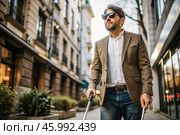 Disability. Loss of vision. A blind man with a cane and dark glasses is walking along a city street. International Day of the Blind on November 13. Стоковое AI-изображение, AI-художник Photojuli / Фотобанк Лори