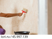 A hand with a wide brush and a bucket next to the wall being repaired during the application of primer or glue before gluing wallpaper. Wall treatment with a primer. Стоковое фото, фотограф Photojuli / Фотобанк Лори