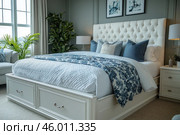 A modern bedroom with a cozy and bright atmosphere. In the center of the room is a large white bed with a soft headboard, decorated with blue and white decorative pillows and a bedspread with a floral pattern. On the sides of the bed are bedside tables with lamps. Green plants in pots are visible on the right and left, which adds freshness and coziness to the interior. On the walls hang paintings in a discreet style, and a large window with light curtains lets in natural light, illuminating the space. Стоковое AI-изображение, AI-художник Анна Кривицкая / Фотобанк Лори