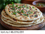 A stack of flatbreads cooked on a griddle or grill. They are garnished with fresh herbs, perhaps cilantro or parsley, adding color to the dish. The flatbreads have a golden, lightly toasted crust with characteristic spots indicating even cooking. The background is complemented by the blurry details of the greenery and perhaps sauces or dips in ceramic bowls, creating a homey or ethnic atmosphere. Стоковое AI-изображение, AI-художник Анна Кривицкая / Фотобанк Лори