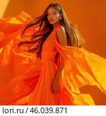 An elegant girl in a long orange dress with fluttering fabric. Her long, silky hair flutters in the wind, creating an effect of dynamics and lightness. The bright orange color of the dress harmonizes with the background, creating an impressive color composition. Makeup emphasizes her expressive features, and gold earrings add a touch of luxury. The atmosphere of the photo conveys sensuality, energy and freedom. Стоковое AI-изображение, AI-художник Анна Кривицкая / Фотобанк Лори