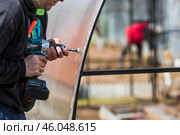 A man holds a battery-powered screwdriver and assembles a new greenhouse made of polyethylene, polycarbonate. Macro photography. Side view. Preparing for the spring garden season. Стоковое фото, фотограф Photojuli / Фотобанк Лори