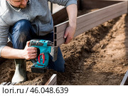 A man holds a battery-powered electric saw and builds new beds in a greenhouse made of wood-polymer composite. Macro photography. Side view. Preparing for the spring garden season. Стоковое фото, фотограф Photojuli / Фотобанк Лори