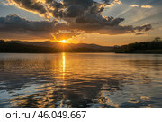 Golden sunset reflecting on calm lake water with dramatic clouds. Стоковое AI-изображение, AI-художник Иван Рябоконь / Фотобанк Лори