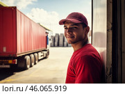 Hispanic truck driver, early 20s, standing near red container truck at busy loading dock. Стоковое AI-изображение, AI-художник Владимир Григорьев / Фотобанк Лори
