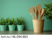 Eco-style composition: in the center is a craft glass filled with wooden cutlery - forks and spoons. The background is painted in a pleasant green color, which harmoniously combines with the surrounding elements. In the background, small pots with green plants are visible, creating a feeling of freshness and a natural theme. The image is made in a minimalist and eco-friendly style, symbolizing conscious consumption and the rejection of plastic in favor of biodegradable materials. Стоковое AI-изображение, AI-художник Анна Кривицкая / Фотобанк Лори