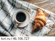 A delicious freshly baked croissant, a white cup of coffee and a cotton napkin on a wooden background. French breakfast. Delicious pastries in close-up. Стоковое AI-изображение, AI-художник Photojuli / Фотобанк Лори