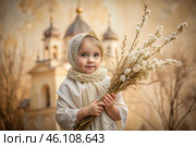 Palm Sunday. Christianity. Portrait of a three-year-old girl in a Russian folk shawl with willow branches in her hands against the background of an Orthodox church. Стоковое AI-изображение, AI-художник Photojuli / Фотобанк Лори