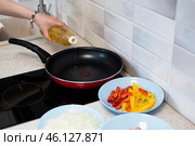 Из бутылки в женской руке льётся растительное масло в разогретую на индукционной плите сковородку, рядом с которой подготовлены нарезанные в тарелках овощи для жарки. Стоковое фото, фотограф Алиса Сокол / Фотобанк Лори