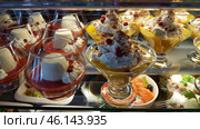 A display case with exquisite desserts in glass goblets. The focus is on two types of desserts: one with panna cotta, decorated with red berry sauce and lingonberries, the other with a delicate mango or citrus mousse, generously decorated with whipped cream, nut crumbles and berries. The desserts look fresh, airy and appetizing. At the bottom of the frame, elements of the appetizers are visible, including salmon and greens, which creates a contrast effect between sweet and salty. Стоковое фото, фотограф Анна Кривицкая / Фотобанк Лори
