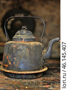 An old metal teapot with sooty walls and peeling paint. It was obviously used to make tea over an open fire, as the bottom is covered in a thick layer of soot and grime. The design of the teapot is simple and functional, with a curved spout and a sturdy metal handle.The background is blurred, but you can see the wooden surface on which the teapot sits, and a partially visible round object resembling a shield. Overall, the image conveys an atmosphere of rustic coziness, camp life, or a medieval kitchen, where similar objects were used in everyday life. Стоковое фото, фотограф Анна Кривицкая / Фотобанк Лори