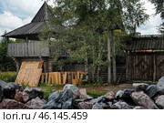 A wooden fortress or fortification built in a traditional style from logs. In the center of the frame, a massive tower with a covered terrace is visible, which can be used as a watchtower or a defensive tower. Along the perimeter, there is a wooden fence with carved gates decorated with complex geometric patterns.In the foreground, building materials are stacked - boards, beams, which may indicate the process of repair or expansion of the building. In the lower part of the frame, piles of stones are visible, possibly used for construction or protection of the territory.The surroundings consist of greenery - trees, bushes and grass, which gives the picture a natural and harmonious atmosphere. Стоковое фото, фотограф Анна Кривицкая / Фотобанк Лори