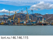Novorossiysk Commercial Sea Port, one of the largest ports in Russia, located on the Black Sea coast. The frame shows cargo ships, port cranes, container yards and part of the industrial infrastructure. In the background are the mountain ranges surrounding the city.Numerous blue and yellow gantry cranes used for loading and unloading ships.Cargo containers stored in the port area.Commercial ships at the berths undergoing cargo operations.Tugboats used to maneuver large ships.Mountains, characteristic of the Novorossiysk landscape.Bright blue sky with clouds, creating a contrast with the industrial landscape. (2023 год). Редакционное фото, фотограф Анна Кривицкая / Фотобанк Лори