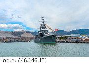 The cruiser Mikhail Kutuzov, permanently moored at the Novorossiysk embankment. This is one of the main museum ships of Russia, which is part of the Central Naval Museum.The cruiser Mikhail Kutuzov is a massive warship with the number "105" on board.It is decorated with signal flags, creating a festive atmosphere.In the background, the port of Novorossiysk, the mountains of the Markotkhsky Range and the berthing facilities are visible.The red naval flag with a white star flutters at the stern.To the right of the ship is the marine terminal, where people and city infrastructure are visible.In the distance, a small tug or boat moves along the water. (2023 год). Редакционное фото, фотограф Анна Кривицкая / Фотобанк Лори