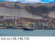 Novorossiysk port against the backdrop of the famous Markotkh ridge. In the foreground is a tugboat, and in the center are three red-and-white chimneys of the Novorossiysk cement plant, one of the oldest enterprises in the region. Numerous port cranes and warehouses are located along the shore.A tugboat in the port waters.An industrial zone with cement plant chimneys.Yellow port cranes unloading ships.The mountains of the Markotkh ridge with their characteristic limestone layers.Industrial buildings and warehouse complexes.Dense vegetation on the mountain slopes. (2023 год). Редакционное фото, фотограф Анна Кривицкая / Фотобанк Лори