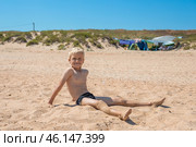 A fair-haired boy sits on the sand by the sea and squints against the bright sun. Tent camp in the background. Summer happy holidays. Стоковое фото, фотограф Коханова Наталья / Фотобанк Лори