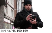 Handsome man wearing black coat and brown leather bag walking down on the city with mobile phone in hand. Stylish bearded middle-aged male using smartphone at urban street. Businessman going on job. Стоковое видео, видеограф Марина Демидюк / Фотобанк Лори