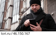 Handsome man wearing black coat and brown leather bag walking down on the city with mobile phone in hand. Stylish bearded middle-aged male using smartphone at urban street. Businessman going on job. Стоковое видео, видеограф Марина Демидюк / Фотобанк Лори