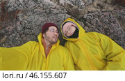 Couple of tourists and bloggers, in love, are lying on rock, wearing cute bright yellow raincoats and warm clothing, during their trip. Стоковое видео, видеограф Strunov David / Фотобанк Лори