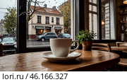 A white cup of hot coffee on a table in a modern cozy cafe by the window. It's raining outside. The concept of comfort and warmth. Стоковое AI-изображение, AI-художник Photojuli / Фотобанк Лори