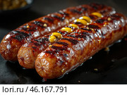 A close-up of grilled sausages with a caramelized crust and grill marks. They are covered in a shiny oily or meaty juice, giving them a delicious look. Mustard has been carefully applied to the surface of one of the sausages.The background is dark and washed out, creating a contrast with the rich color of the meat. This image perfectly captures the atmosphere of a barbecue, picnic, or open-fire grilling. Стоковое AI-изображение, AI-художник Анна Кривицкая / Фотобанк Лори