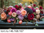 An elegant floral arrangement adorned with roses, dahlias, ranunculus, berries and various greens. The bouquet is made in deep shades of burgundy, lilac, pink and peach, creating an atmosphere of sophistication and luxury. The flowers are arranged on a black lacquered object with decorative fittings, which gives the scene a mournful character. The background is slightly blurred, with autumnal notes of nature, adding depth and emotional shade to the composition. The atmosphere of the photo conveys grief, memory and respect. Стоковое AI-изображение, AI-художник Анна Кривицкая / Фотобанк Лори