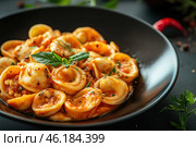A delicious pasta dish, probably tortellini or orecchiette, served in a deep black bowl. The pasta is covered in a thick, spiced tomato sauce, perhaps with chunks of meat or cheese. The dish is garnished with fresh green basil leaves and sprinkled with ground black pepper and chopped herbs. Small sprigs of parsley, hot red peppers and seasonings are scattered around the plate, creating a gourmet presentation. The atmosphere of the photo is warm and cozy, with an emphasis on the rich colors of Italian cuisine. Стоковое AI-изображение, AI-художник Анна Кривицкая / Фотобанк Лори