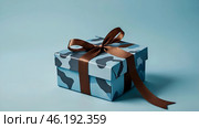 The concept of Defender of the Fatherland Day on February 23. Gift camouflage box with a festive satin ribbon bow on a light blue background. Стоковое AI-изображение, AI-художник Photojuli / Фотобанк Лори