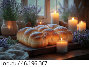 A cozy home scene with fragrant pastries beautifully served on a wooden board. These are fresh, soft, airy buns with a golden crust and a light dusting of flour or powdered sugar. They are placed on a rustic table surrounded by candles that create a warm and romantic atmosphere. The frame also includes lavender bouquets in glass and burlap vases, and soft daylight is visible through the window, complementing the cozy atmosphere. Стоковое AI-изображение, AI-художник Анна Кривицкая / Фотобанк Лори