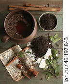 Still life with brewed tea in a clay mug, loose black tea, green tea plant leaves and cinnamon sticks. The surface of the wooden table is aged and covered with green paint, which adds a vintage character to the composition. Scattered old book pages, a ceramic vessel and spices create an atmosphere of coziness, tradition and naturalness. Стоковое AI-изображение, AI-художник Анна Кривицкая / Фотобанк Лори