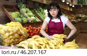 Positive young asian salesgirl standing next to produce stand in fruit and vegetable section of supermarket, laying out ripe bananas for sale. Стоковое видео, видеограф Яков Филимонов / Фотобанк Лори